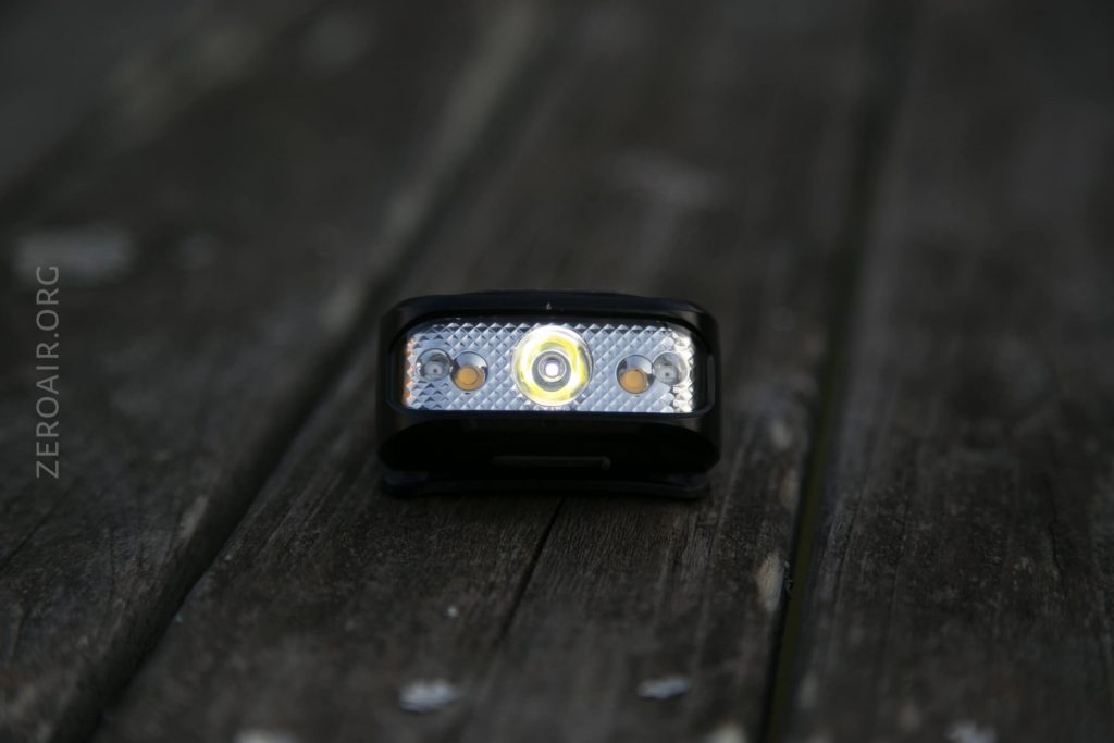 A small rectangular LED headlamp with three bulbs is placed on a weathered wooden surface. The main bulb is centered, with two smaller bulbs on either side. The background is blurred.