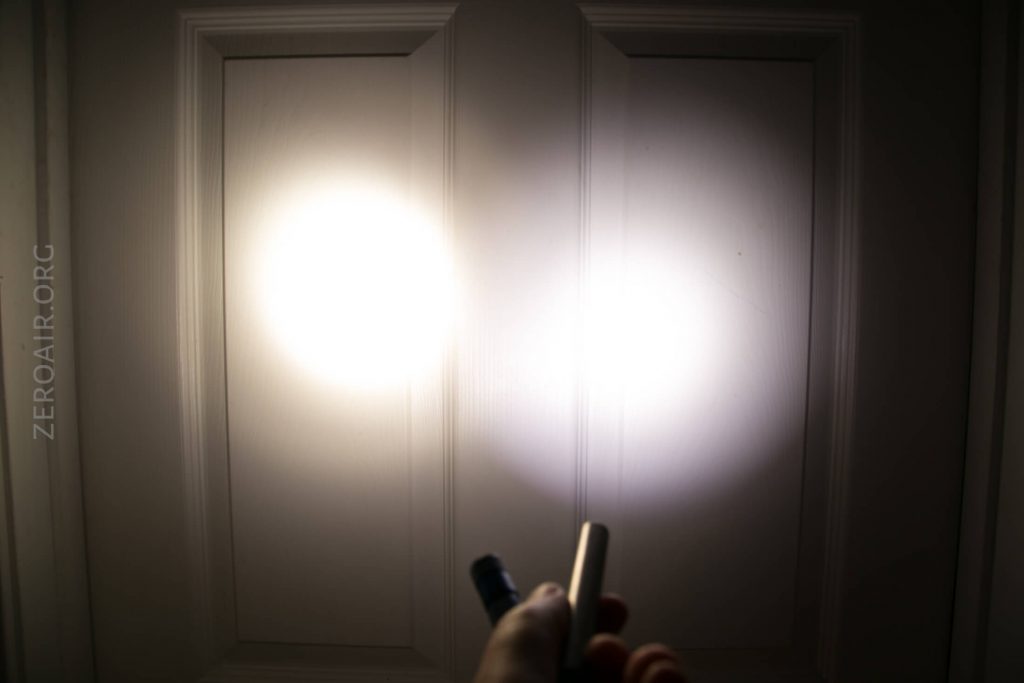A hand holds two flashlights, shining their beams next to each other on a white paneled door in a dim setting. The left beam appears larger and warmer; the right beam is smaller and cooler in tone.