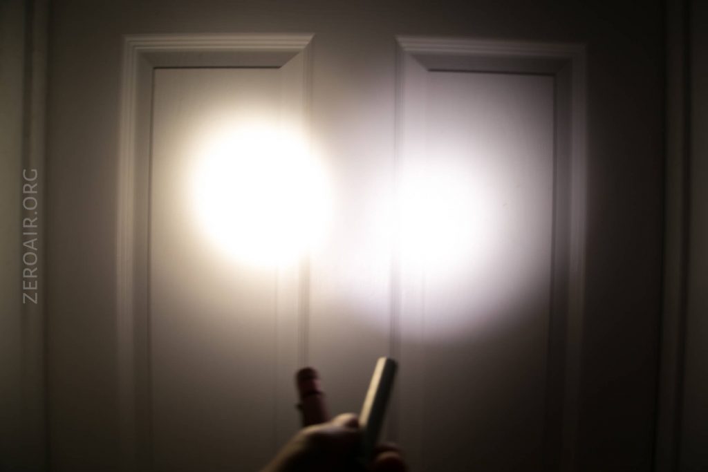 A person holds a flashlight pointed at a white, paneled door, creating two overlapping circles of bright light on the doors surface. The website ZEROAIR.ORG is faintly visible on the left side.