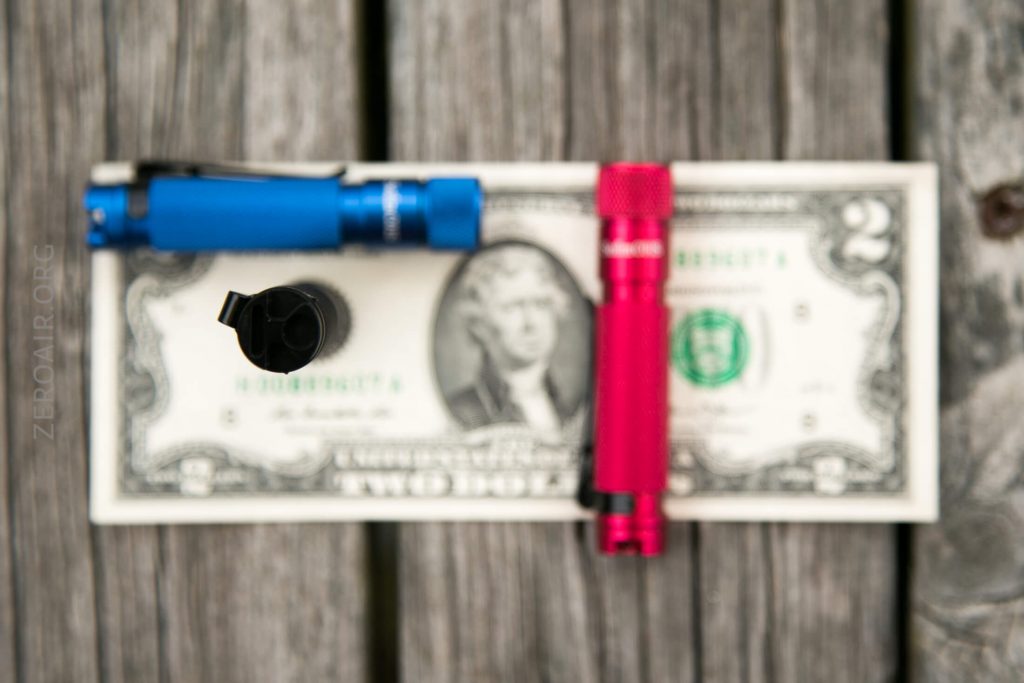 A blue flashlight, a black item, and a red flashlight are placed on a two-dollar bill on a wooden surface; the image is out of focus.