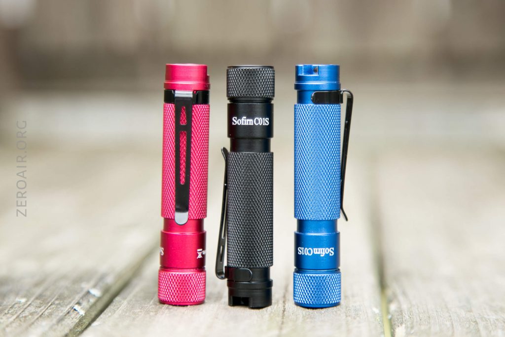 Three compact flashlights—one red, one black, and one blue—stand upright on a wooden surface. Each has a textured grip, a clip, and the text “Sofirn C01S” is visible on the black and blue flashlights.