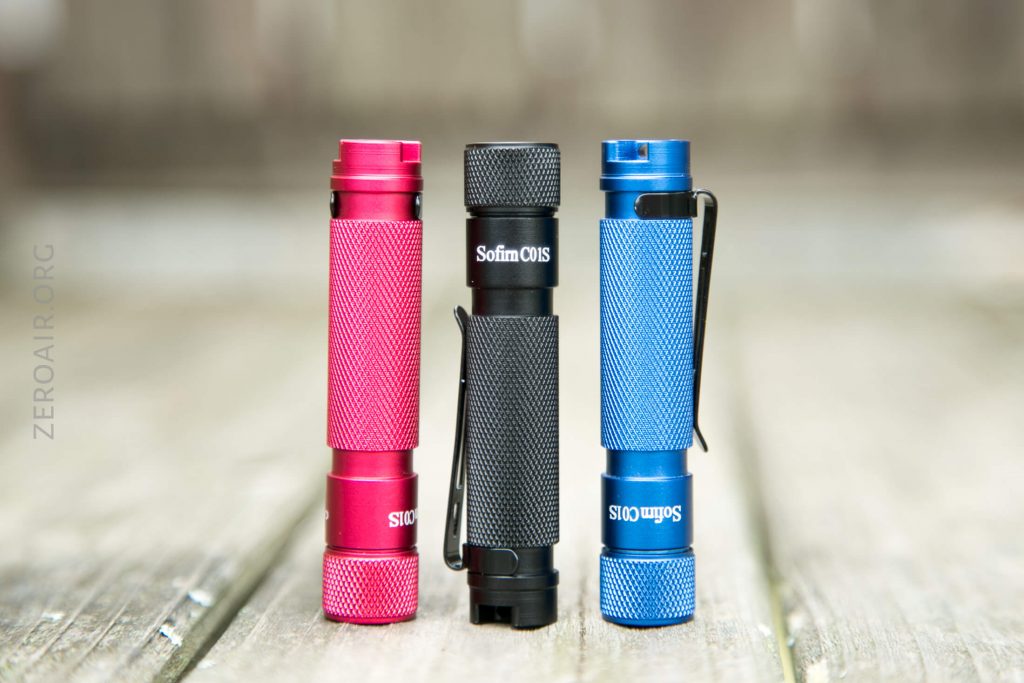 Three compact Sofirn flashlights in red, black, and blue stand upright on a wooden surface, each featuring a textured grip and metal clip, with a blurred background.