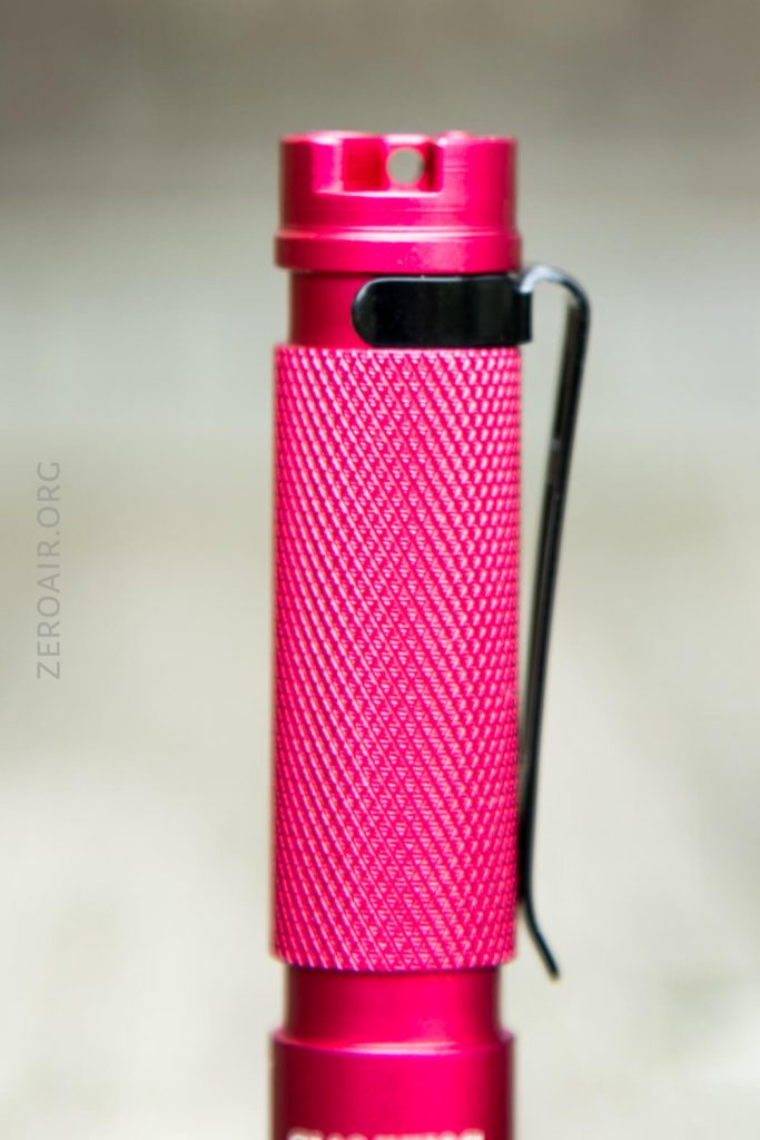 A close-up image of a small red flashlight with a textured grip and a black pocket clip, shown vertically against a blurred neutral background.