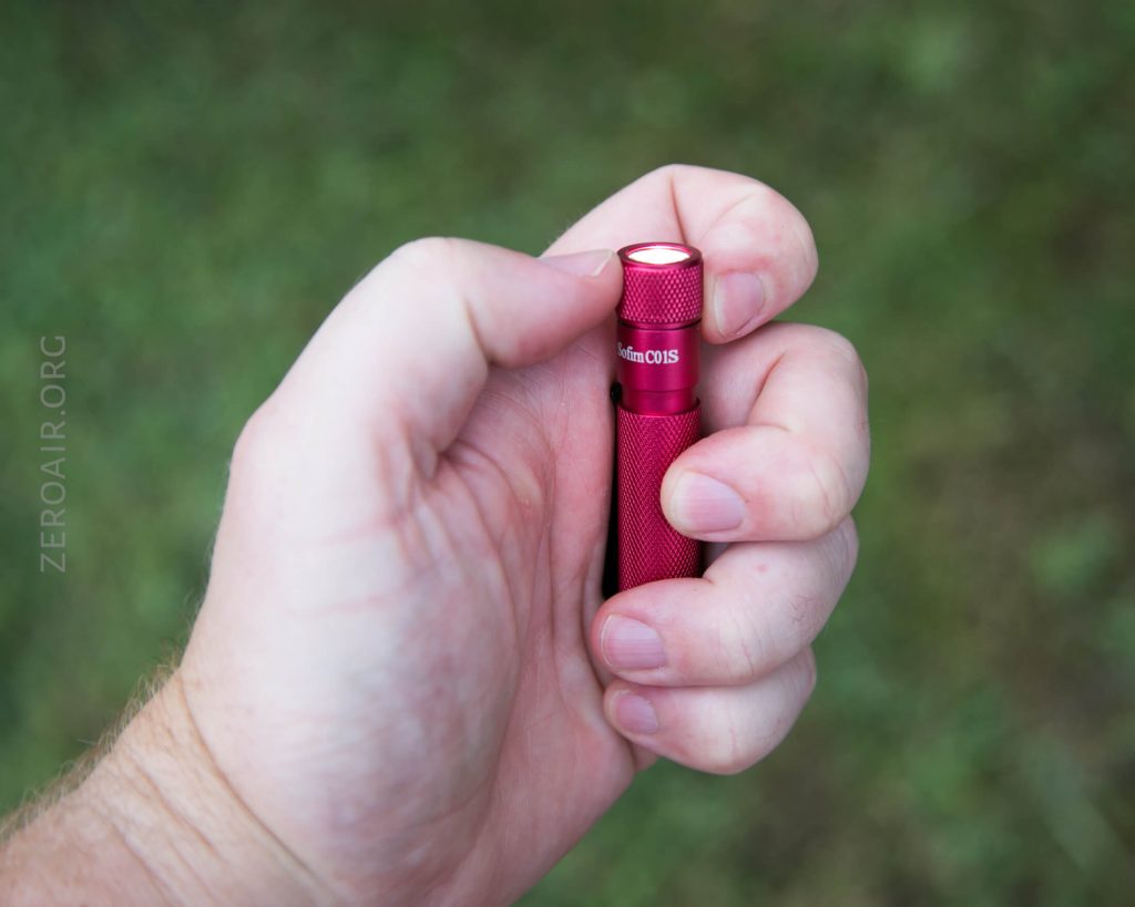 A hand holding a small red flashlight with the thumb positioned on the button at the end. The background is grassy and out of focus.