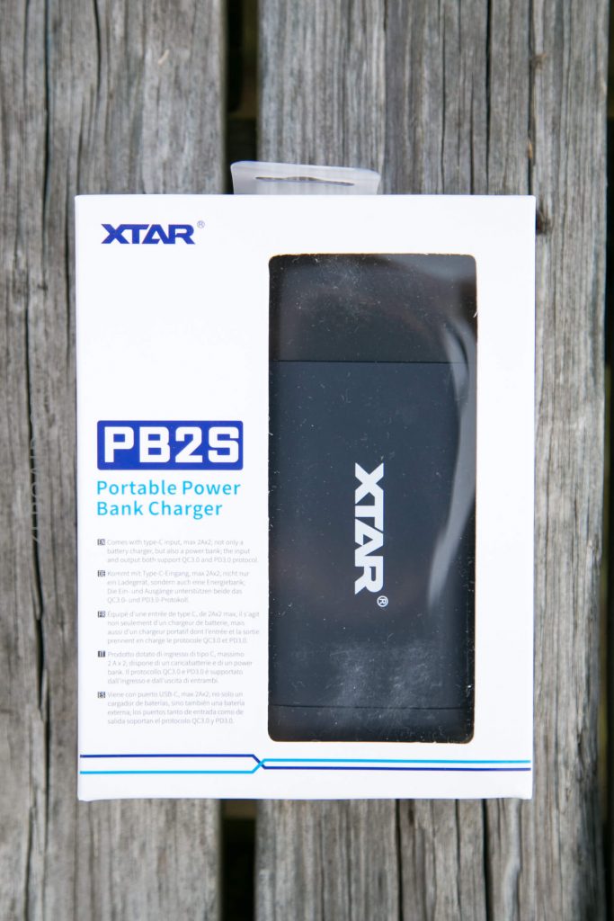 A boxed XTAR PB2S portable power bank charger is displayed on a weathered wooden surface. The charger is visible through a transparent window on the front of the white packaging.