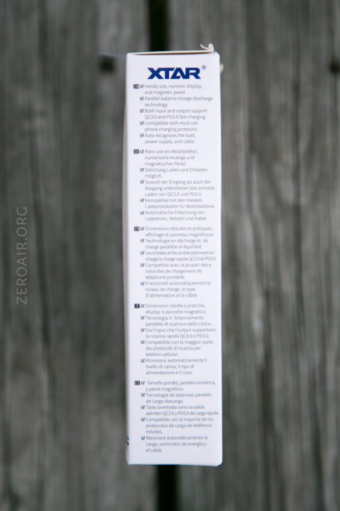 A close-up of the side of an XTAR product box listing features in multiple languages. The text is printed in columns on a white background, with a blurred wooden surface in the background.