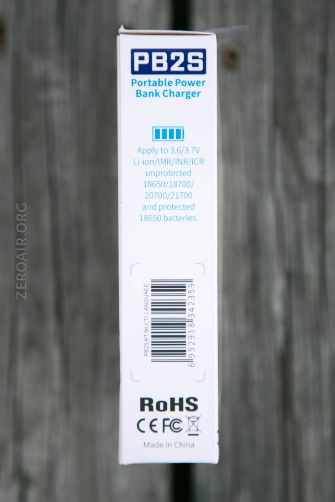 White product packaging for the PB2S Portable Power Bank Charger, showing specifications for compatible Li-ion batteries, a barcode, RoHS and CE marks, and “Made in China” text. Background is gray wood.