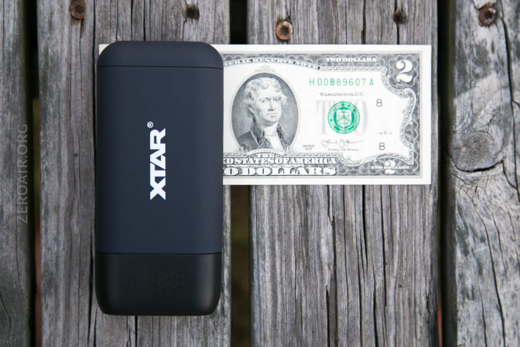 A black XTAR device is placed next to a U.S. two-dollar bill on a weathered wooden surface, showing a size comparison between the device and the bill.