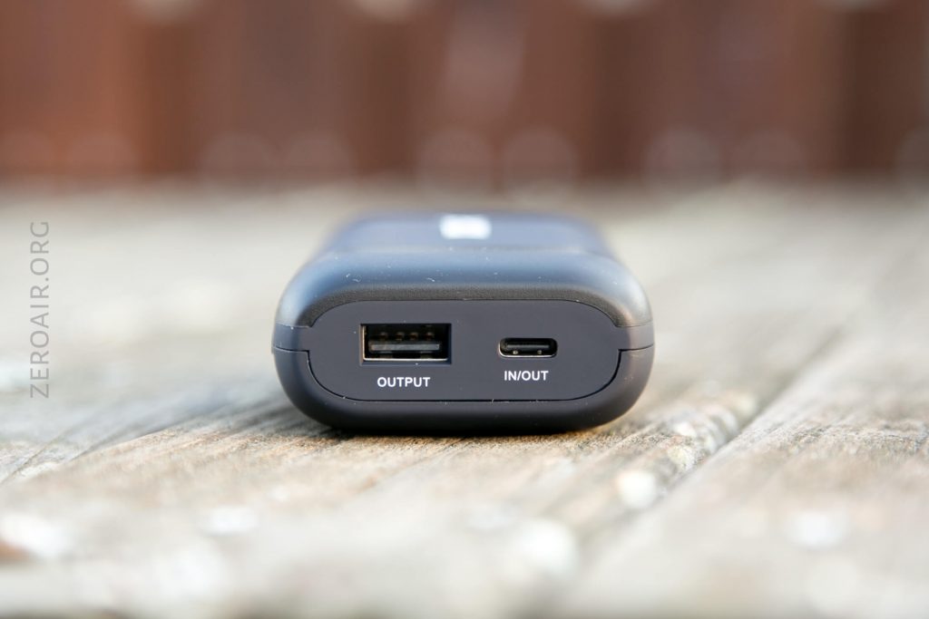 A close-up of a black device showing a USB-A output port labeled OUTPUT and a USB-C port labeled IN/OUT, placed on a wooden surface.
