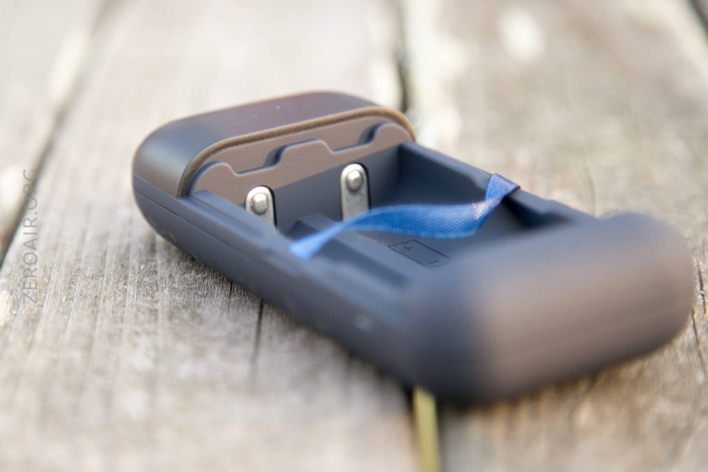 A close-up of an empty black battery charger with metal contacts and a blue ribbon inside, resting on a weathered wooden surface.
