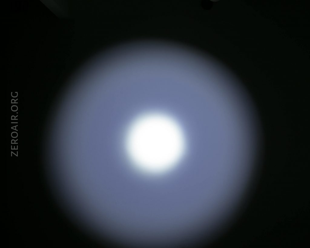 A circular white light beam with a bright center and a softer blueish outer ring is projected onto a dark surface. The text ZEROAIR.ORG is visible on the left side of the image.