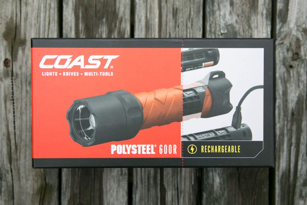 A Coast Polysteel 600R rechargeable flashlight is shown in its packaging, which features product images and text highlighting that it is rechargeable. The box rests on a wooden surface.