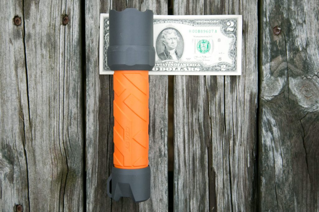 An orange and gray flashlight is placed horizontally on top of a two-dollar bill, both lying on a weathered wooden surface. The flashlight appears slightly longer than the bill.