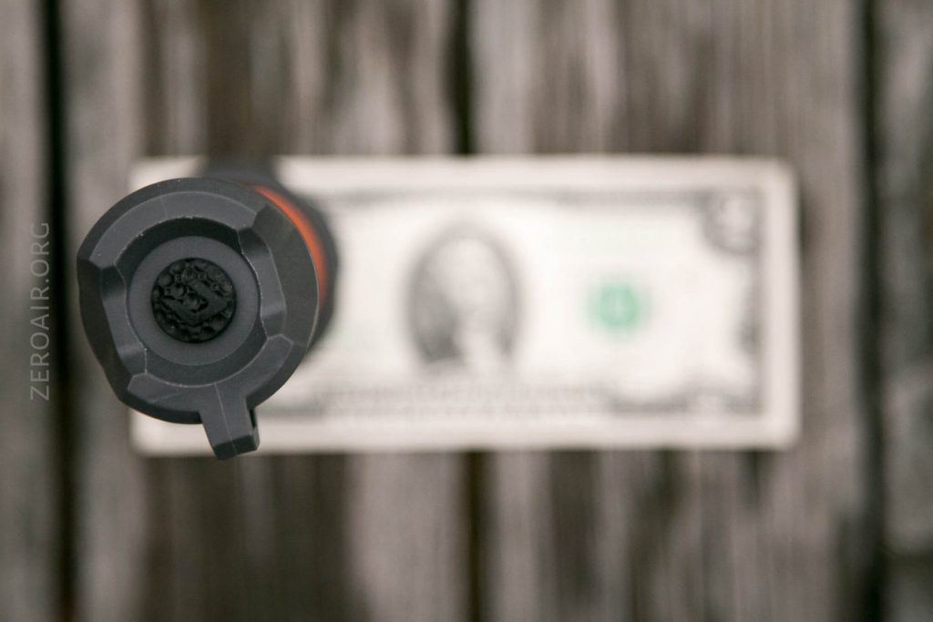 A handgun pointed downward is in focus in the foreground, with a blurred one-dollar bill lying flat on a wooden surface below it.