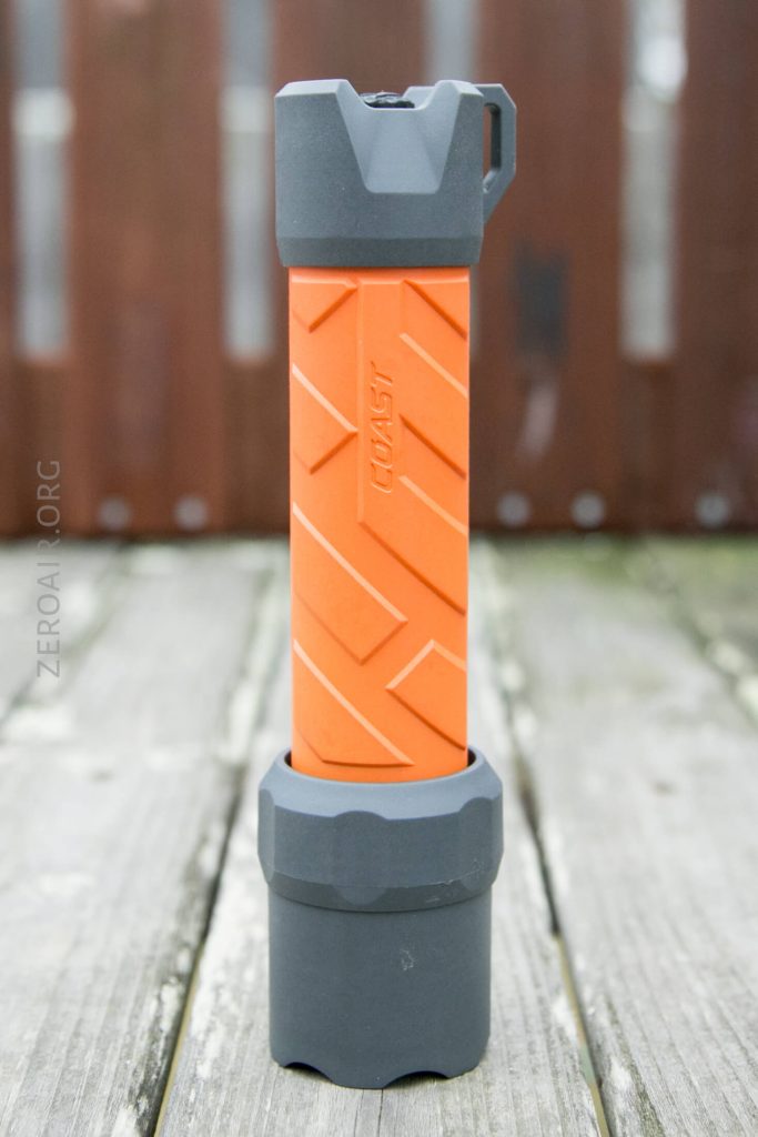 An orange and gray flashlight stands upright on a weathered wooden surface with a blurred brown fence in the background. The flashlight has textured grip patterns and a brand logo on the side.