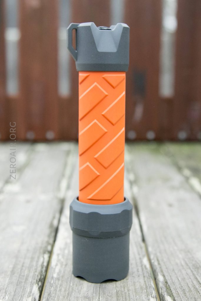 A flashlight with a bright orange textured grip and dark gray ends stands upright on a wooden surface, with a blurred brown fence in the background.