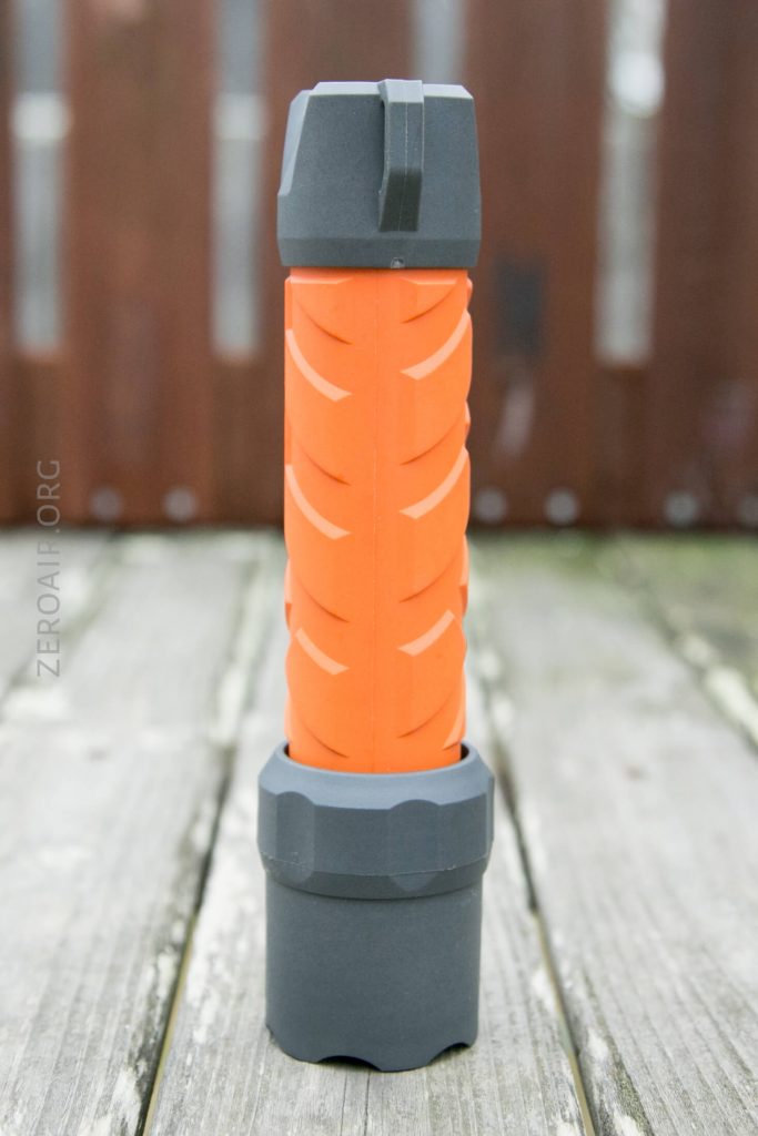 A cylindrical flashlight with a textured orange grip and dark gray ends is standing upright on a weathered wooden surface outdoors.