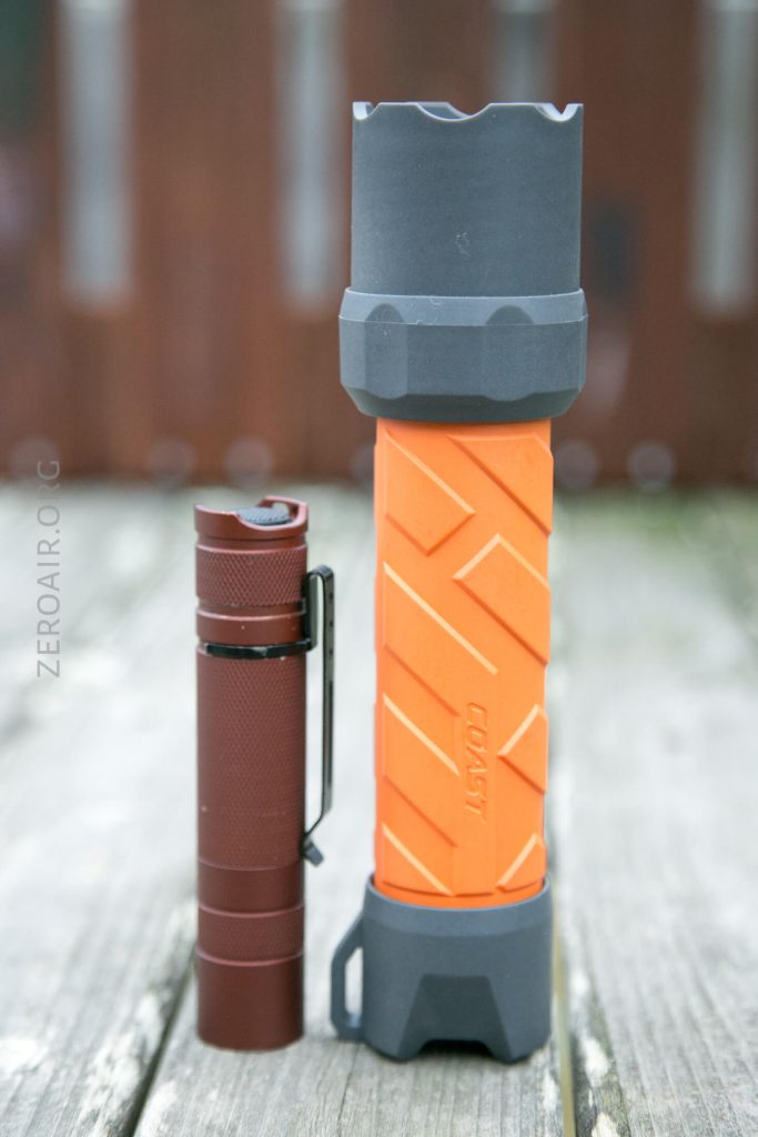 A small red flashlight stands upright next to a larger, orange and grey flashlight on a wooden surface with a blurred background.