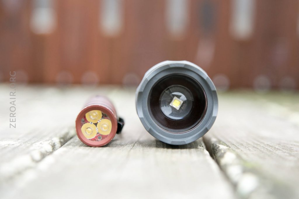 A flashlight is shown disassembled on a wooden surface. The head, with a single LED visible inside, is on the right, and the body, displaying three battery contacts, is on the left.