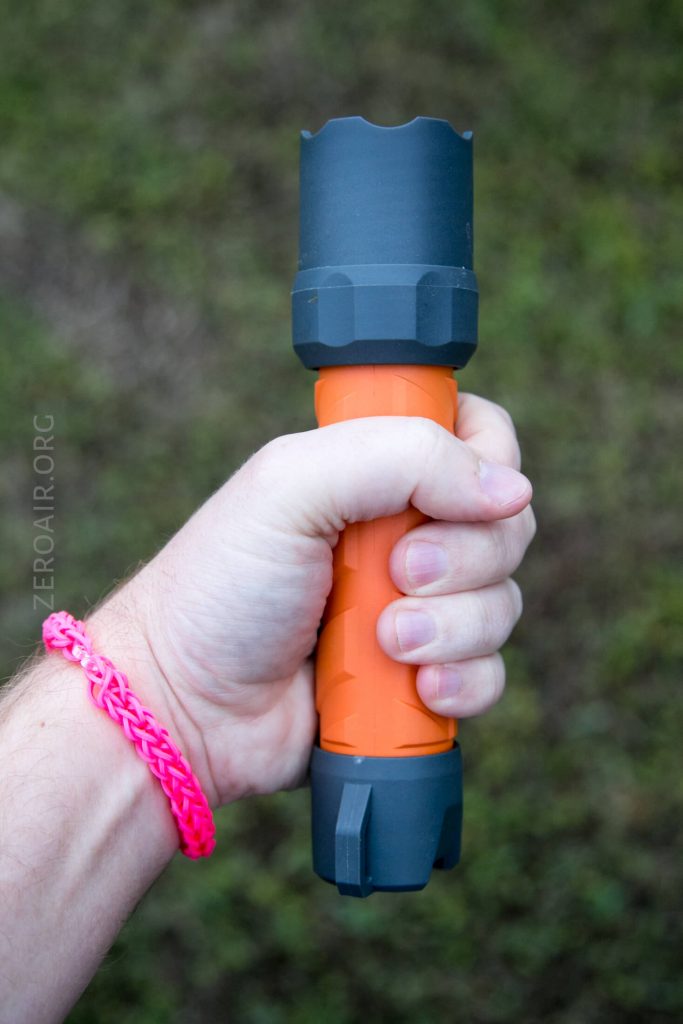 A hand holding an orange and gray flashlight outdoors, with the person wearing a pink woven bracelet on their wrist. The background is out of focus grass.