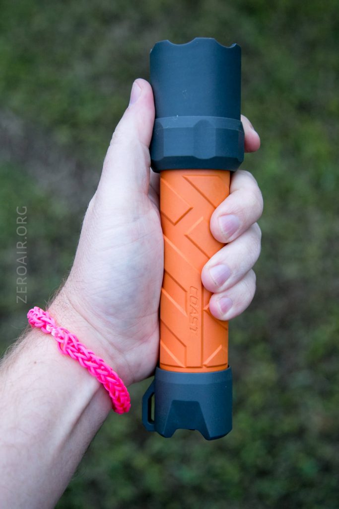 A person wearing a pink bracelet holds an orange and black flashlight with a textured grip. The background is blurred grass. The word COAST is visible on the flashlight.