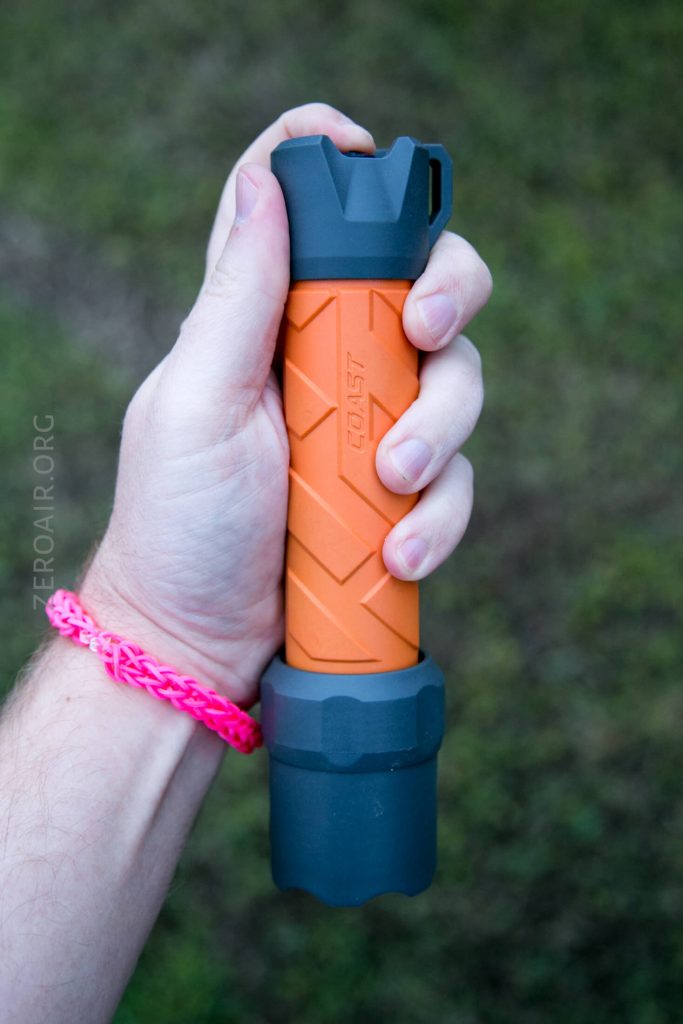 A hand holding an orange and gray flashlight with a textured grip, photographed outdoors against a blurred green background. The wrist has a pink woven bracelet.