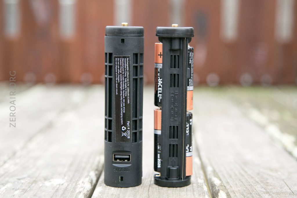 Two black cylindrical battery holders are standing upright on a wooden surface. One holder is empty, showing its internal structure, while the other contains four AA batteries. A USB port is visible at the bottom of the empty holder.