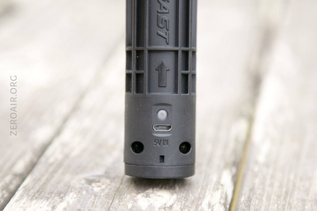 Close-up of the bottom part of a black cylindrical device with a micro USB charging port labeled 5V IN, a small button, and an upward-pointing arrow, placed on a wooden surface.