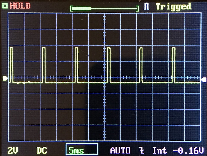 Oscilloscope display showing a repeating square wave signal with regular pulses, yellow trace on a grid. The screen displays settings including 2V, DC, 5ms, AUTO, and a voltage value of -0.16V.