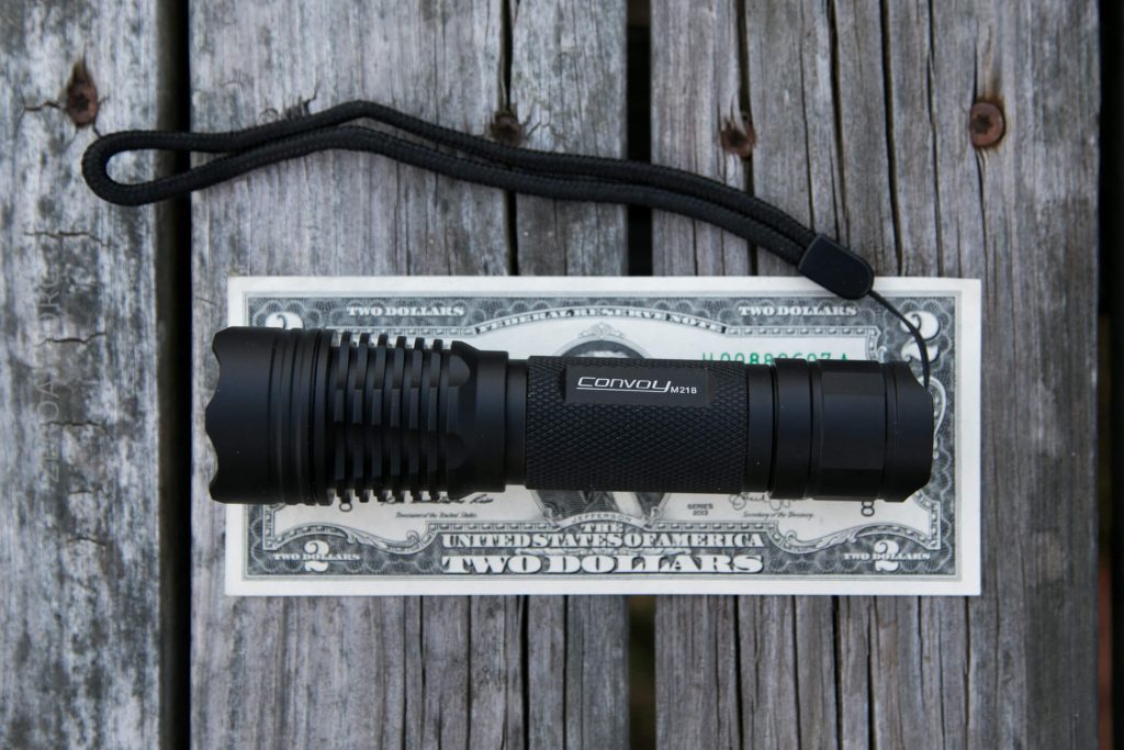 A black flashlight with a wrist strap is placed on top of a two-dollar bill, both resting on a weathered wooden surface.