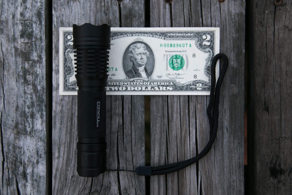 A black flashlight with a wrist strap is placed on a weathered wooden surface, positioned in front of a US two-dollar bill for size comparison.