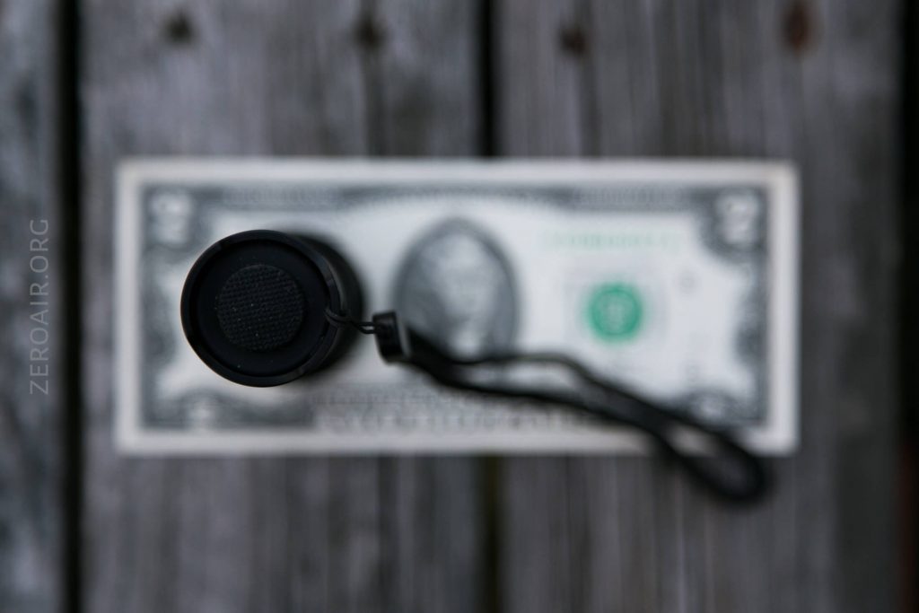 A small cylindrical flashlight with a wrist strap is positioned vertically on top of a one-dollar bill, both resting on a weathered wooden surface. The image is focused on the flashlight, with the bill slightly out of focus.