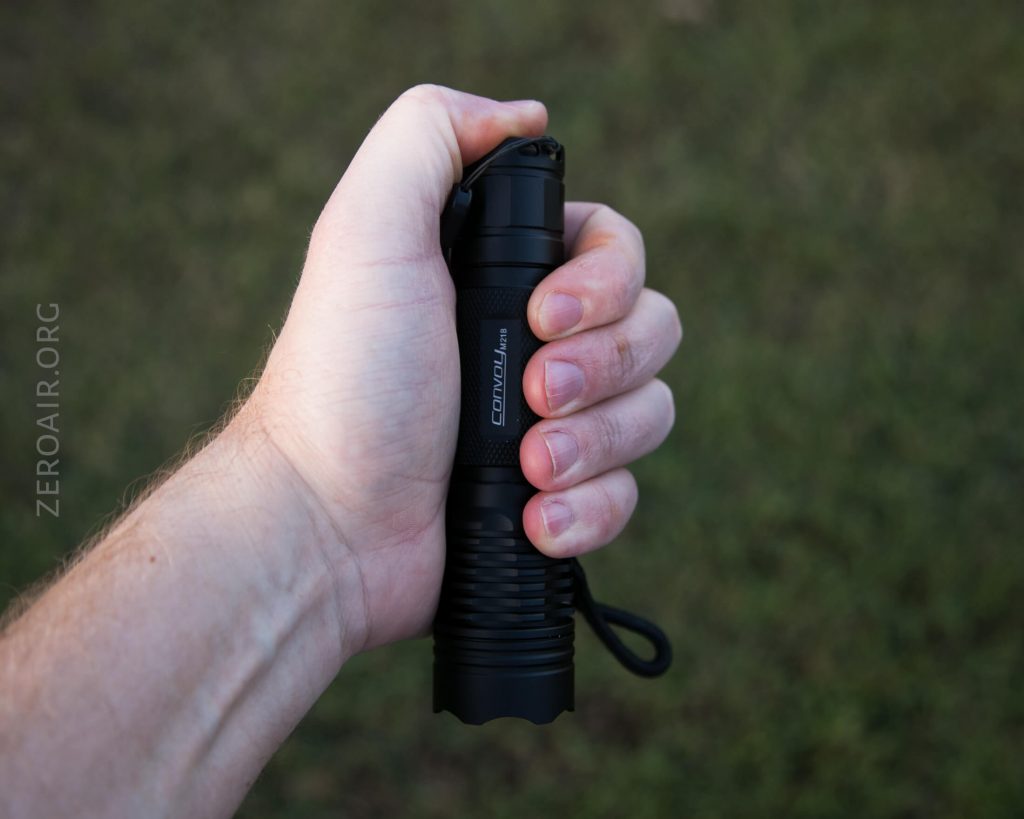 A person holding a black Convoy flashlight in their right hand, with a grassy background and a wrist strap attached to the flashlight.