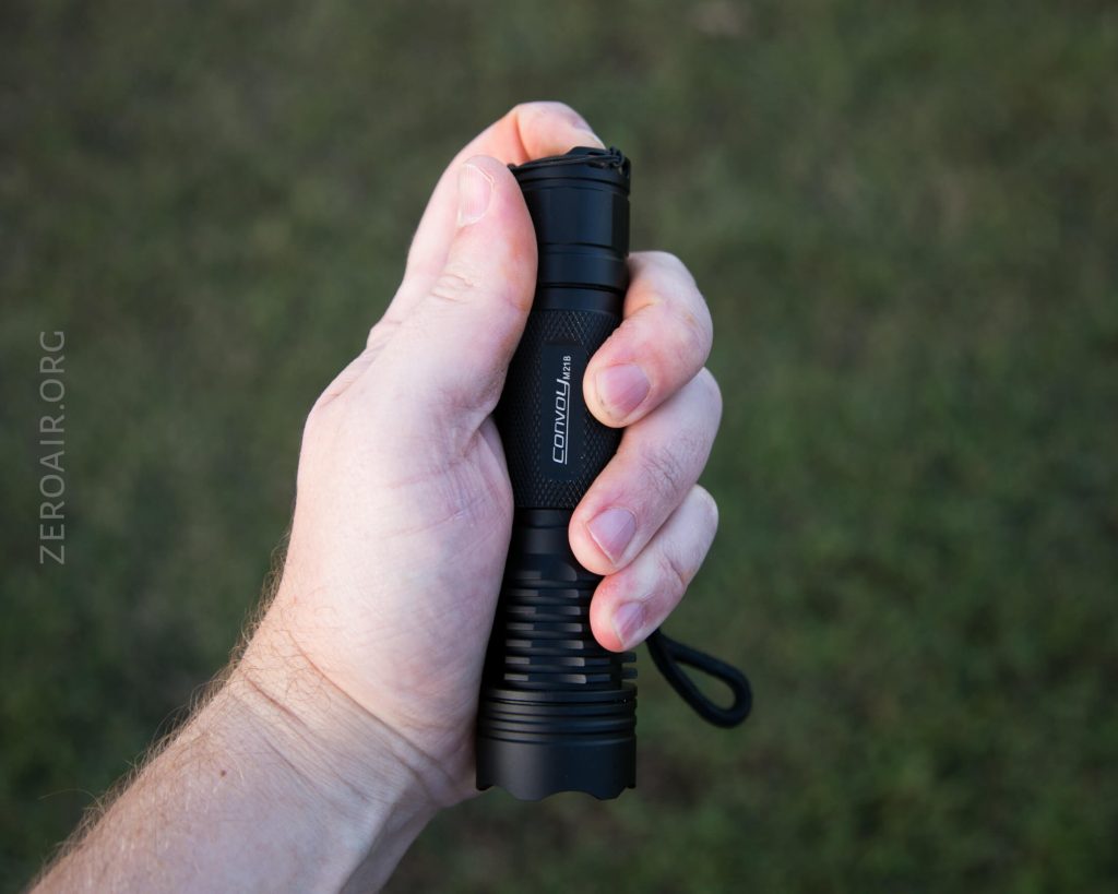 A person holding a black Convoy flashlight in their right hand, with a grassy background visible out of focus.