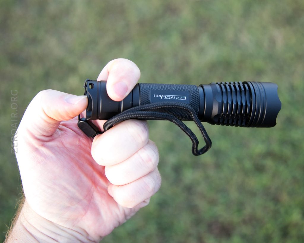 A hand holding a black Convoy flashlight with a wrist strap, positioned against an outdoor background of green grass.