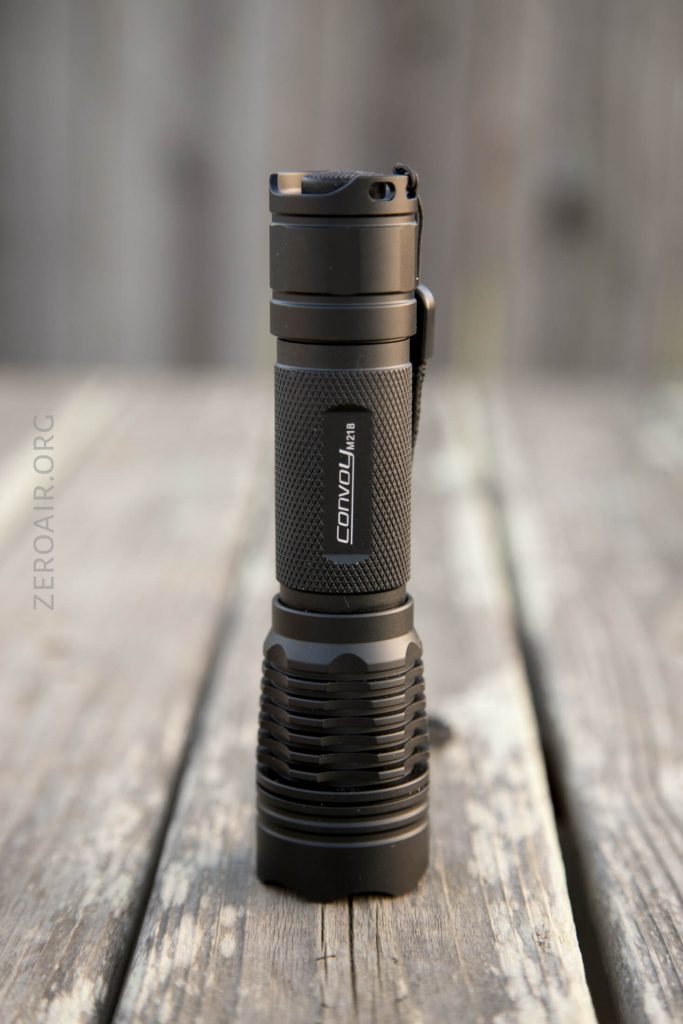 A black Convoy flashlight stands upright on a weathered wooden surface with a blurred background. The flashlight features a textured grip and finned heat dissipation design.