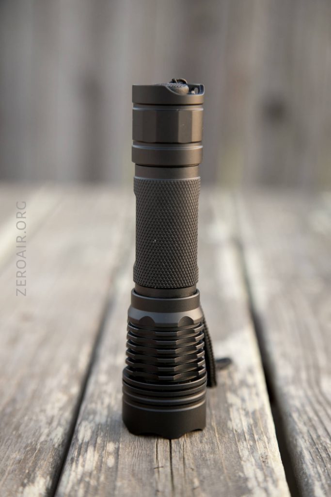 A black, cylindrical flashlight with a textured grip stands upright on a weathered wooden surface. The background is blurred, drawing attention to the flashlight.