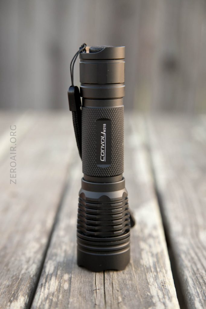 A black Convoy M3 flashlight stands upright on a weathered wooden surface, with a lanyard attached to the top and the brand name visible on the body. The background is blurred.
