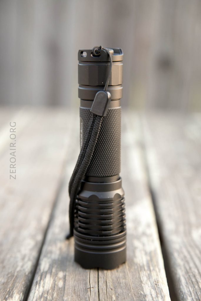 A black tactical flashlight with a textured grip, metal clip, and wrist strap stands upright on a weathered wooden surface. The background is blurred wood planks.