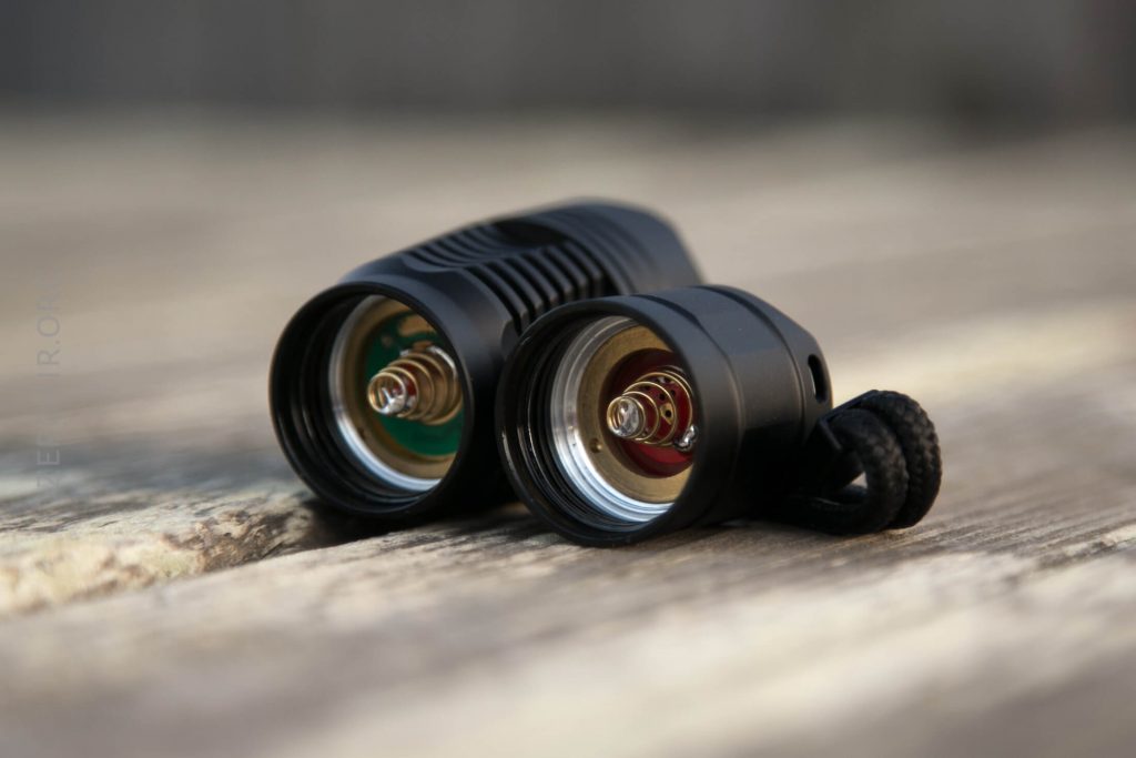 Two black flashlight bodies lie side by side on a wooden surface, both with their tailcaps removed to show the battery contacts and springs inside. One has a wrist strap attached.