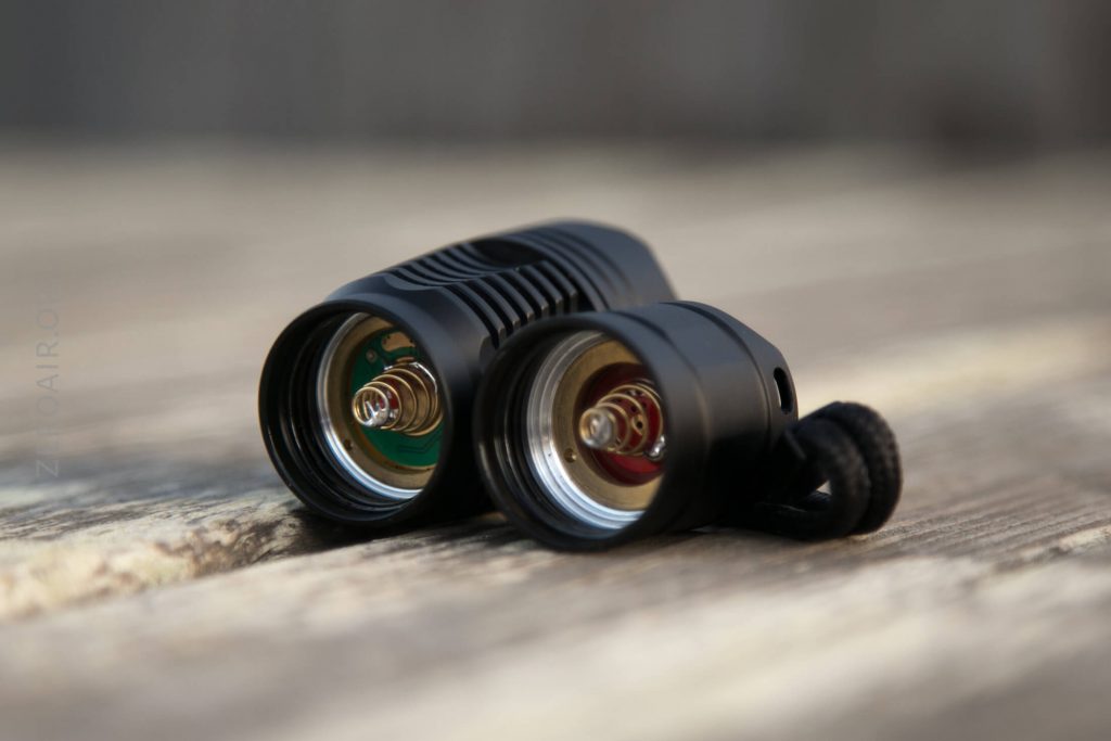 A close-up of a black flashlight lying on its side with the battery compartment open, exposing the metal contacts inside. The flashlight rests on a wooden surface.