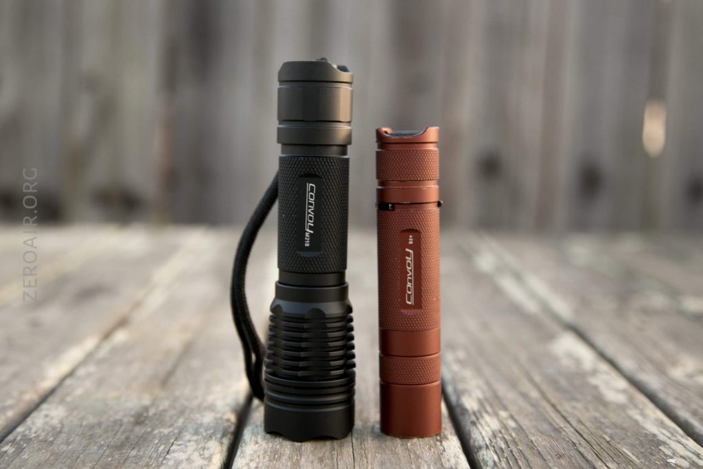 Two flashlights stand upright on a wooden surface. The left flashlight is black with a textured grip and lanyard, and the right flashlight is red with a smooth, slim design. A blurred wooden fence is in the background.