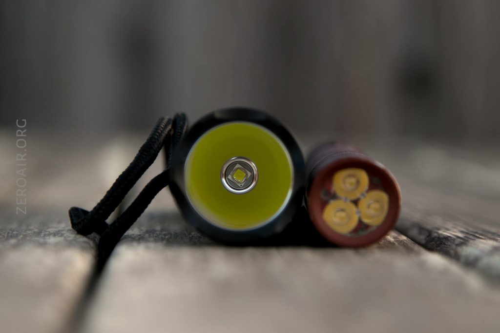 A close-up view of the front of a flashlight next to the bottom of a battery pack with four circular contact points, both lying on a wooden surface.