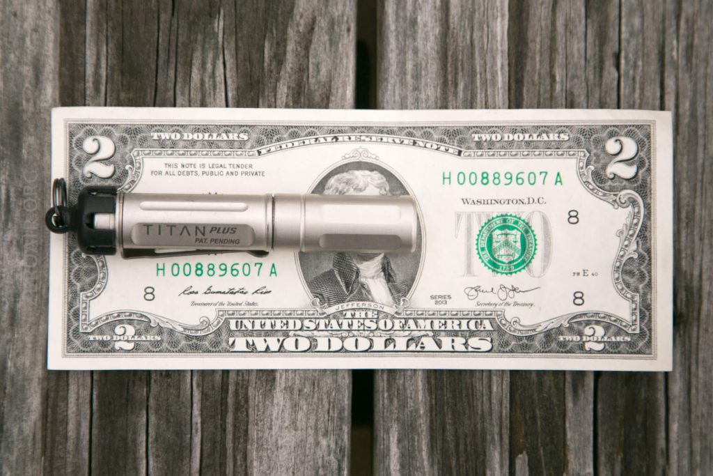 A compact silver flashlight labeled TITAN PLUS is placed on top of a two-dollar bill, which is lying on a wooden surface.