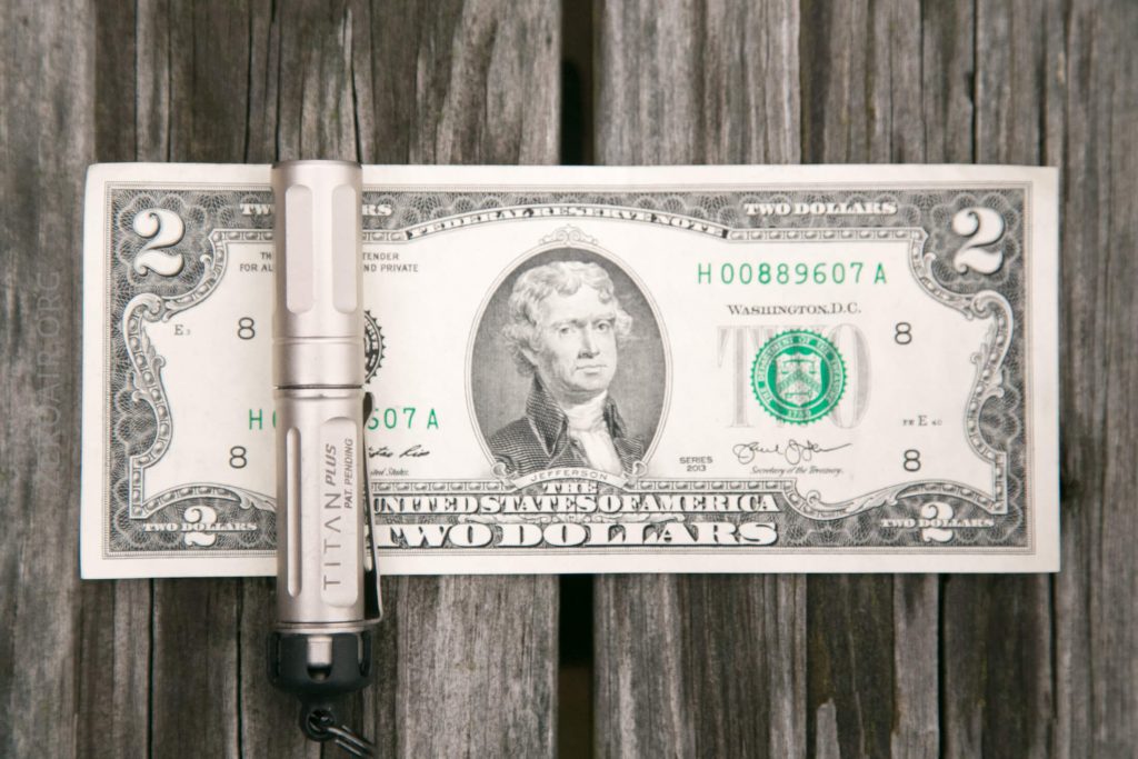A small metal flashlight labeled TITAN rests horizontally across the left side of a U.S. two-dollar bill, which is placed on a weathered wooden surface.