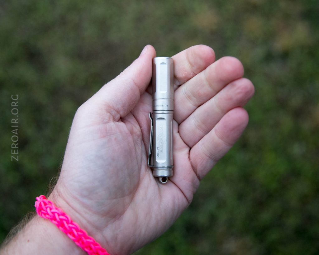 A hand holds a small silver flashlight against a blurred outdoor background. The person wears a pink braided bracelet on their wrist. The text ZEROAIR.ORG is visible on the left side of the image.