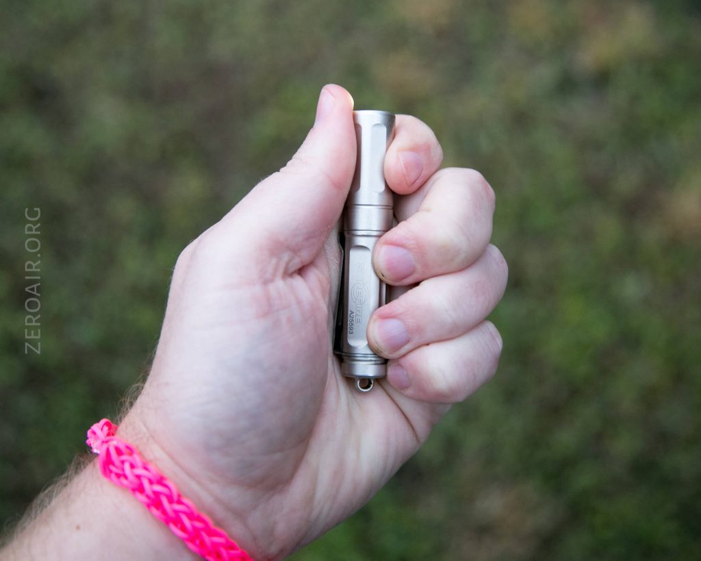 A person holding a small metallic flashlight in their right hand, with a pink woven bracelet on their wrist. The background is out of focus grass. The text ZEROAIR.ORG is visible along the left edge.