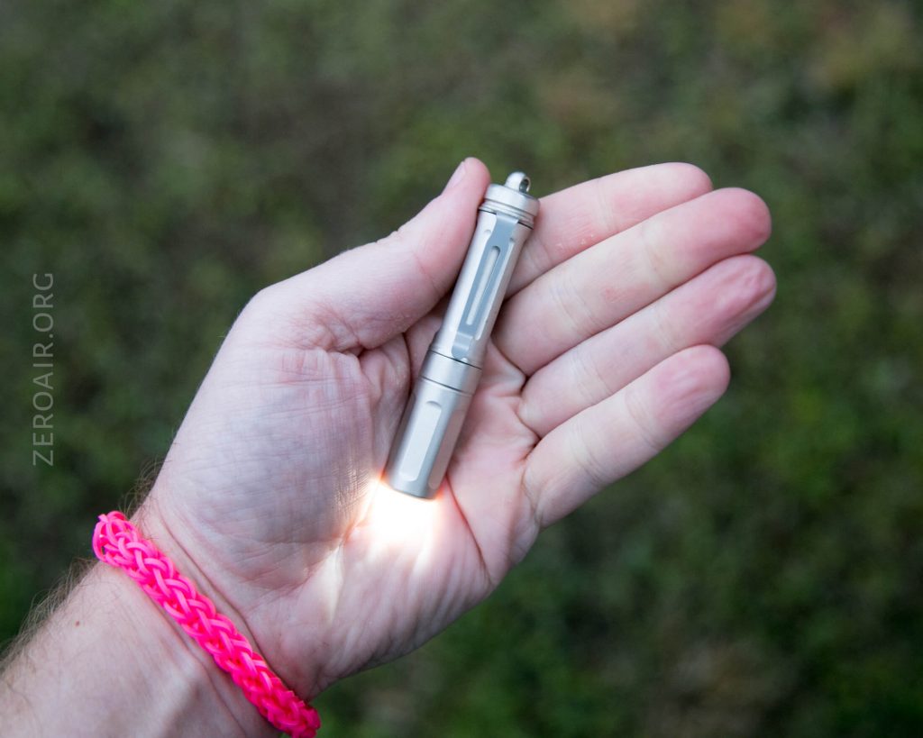 A hand holding a small, silver flashlight turned on, emitting light. The person is wearing a bright pink woven bracelet, and the background is blurred green grass. The text ZEROAIR.ORG appears on the left side.