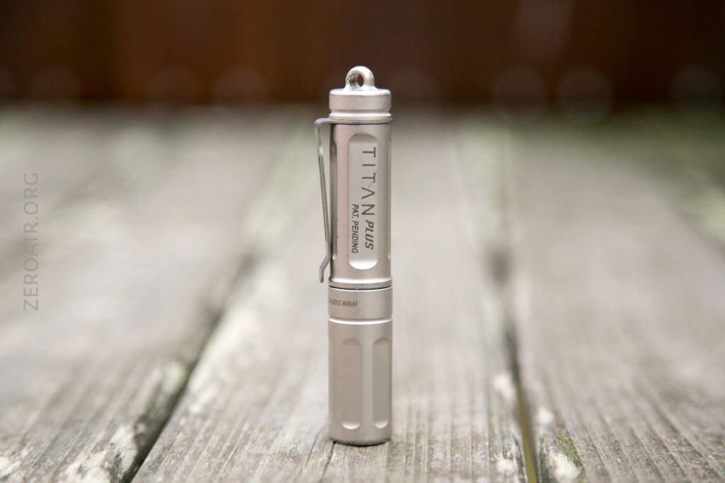 A small, silver flashlight labeled Titan Plus stands upright on a weathered wooden surface. The flashlight has a clip and a keyring attachment at the top.