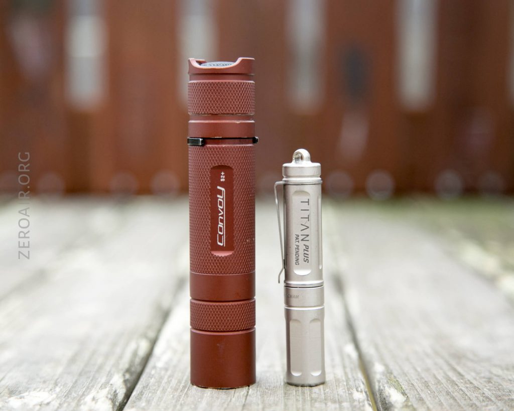 A brown Convoy S2+ flashlight and a smaller silver Maratac Titanium flashlight stand upright on a weathered wooden surface with a blurred outdoor background.